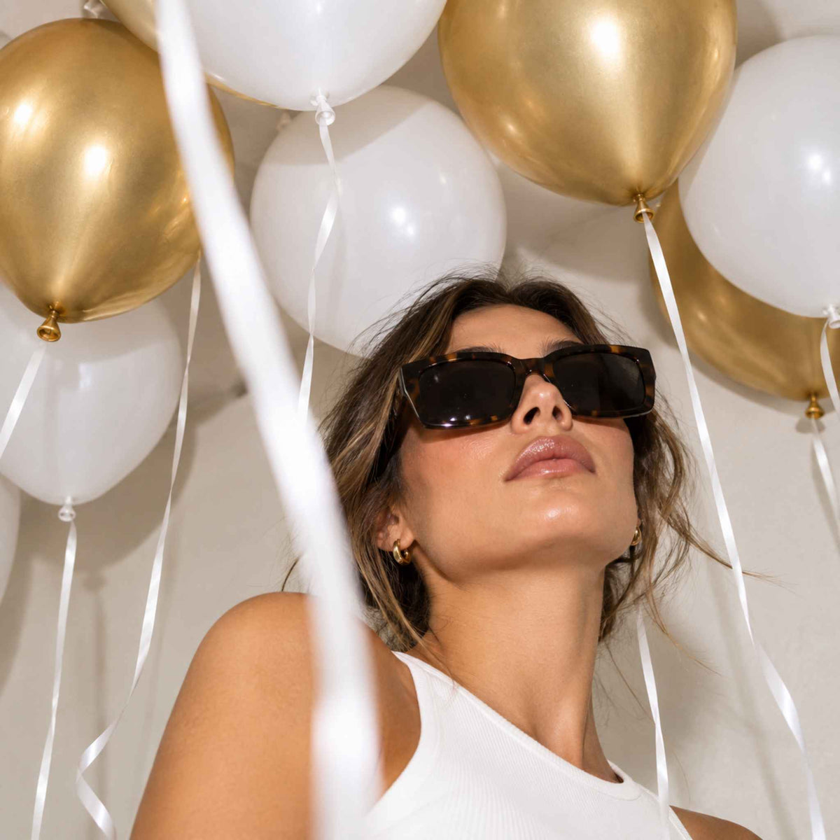 A woman wearing dark brown rectangular sunglasses and a white ribbed top, looking upward among white and gold balloons with white ribbons.