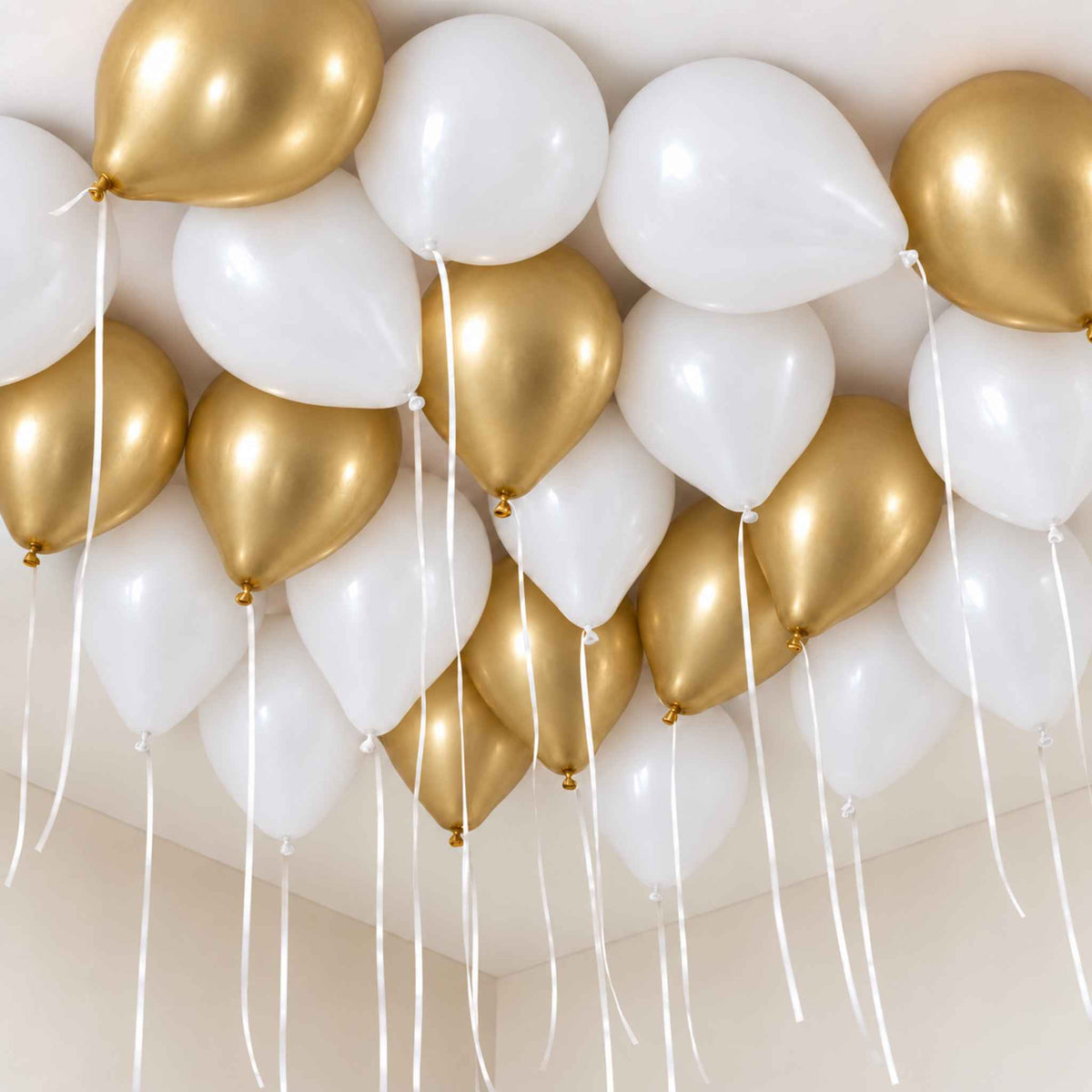 A cluster of metallic gold and matte white balloons arranged against a light beige wall and ceiling, with long white ribbons hanging down.