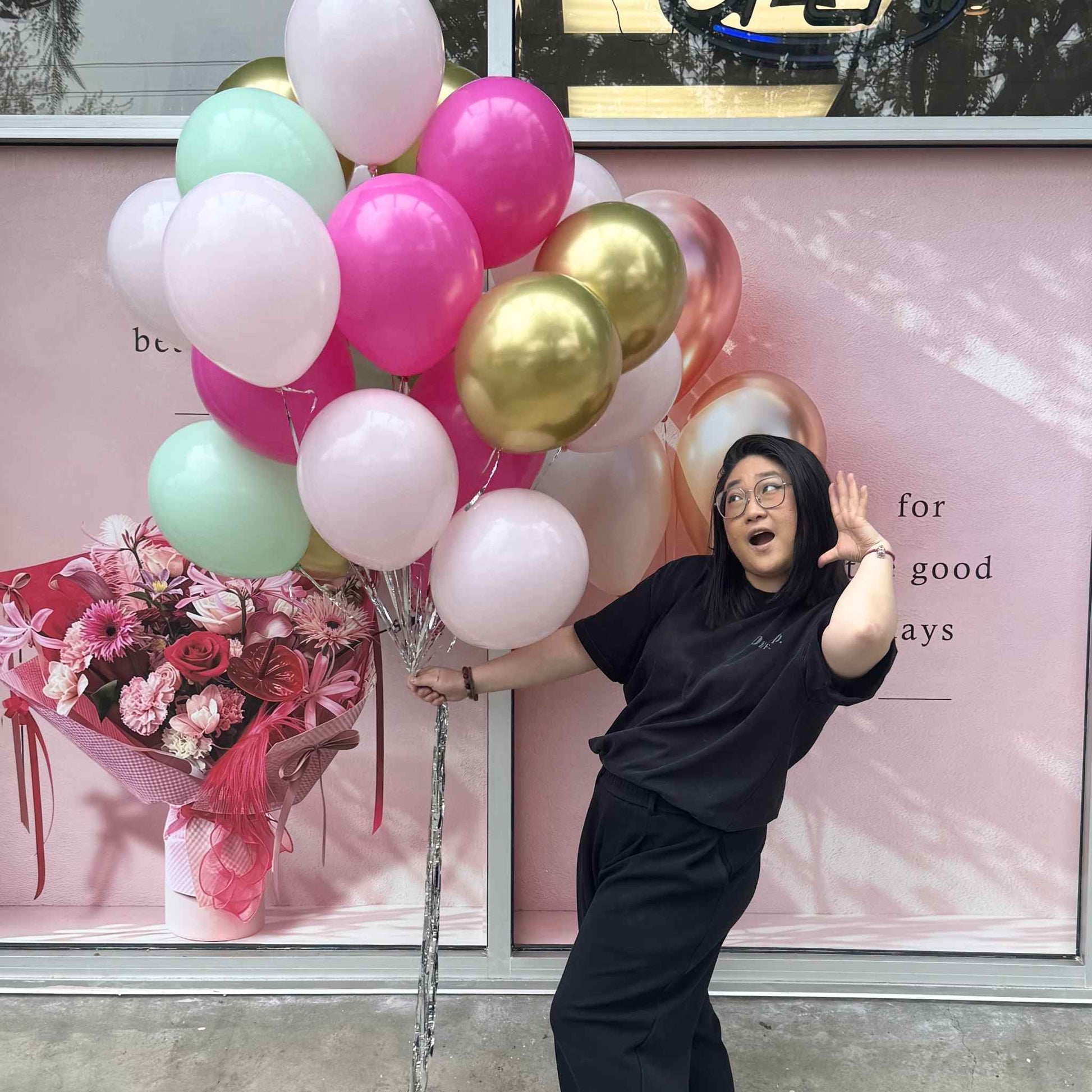 Person holding balloons and flowers in front of a store window with promotional posters.