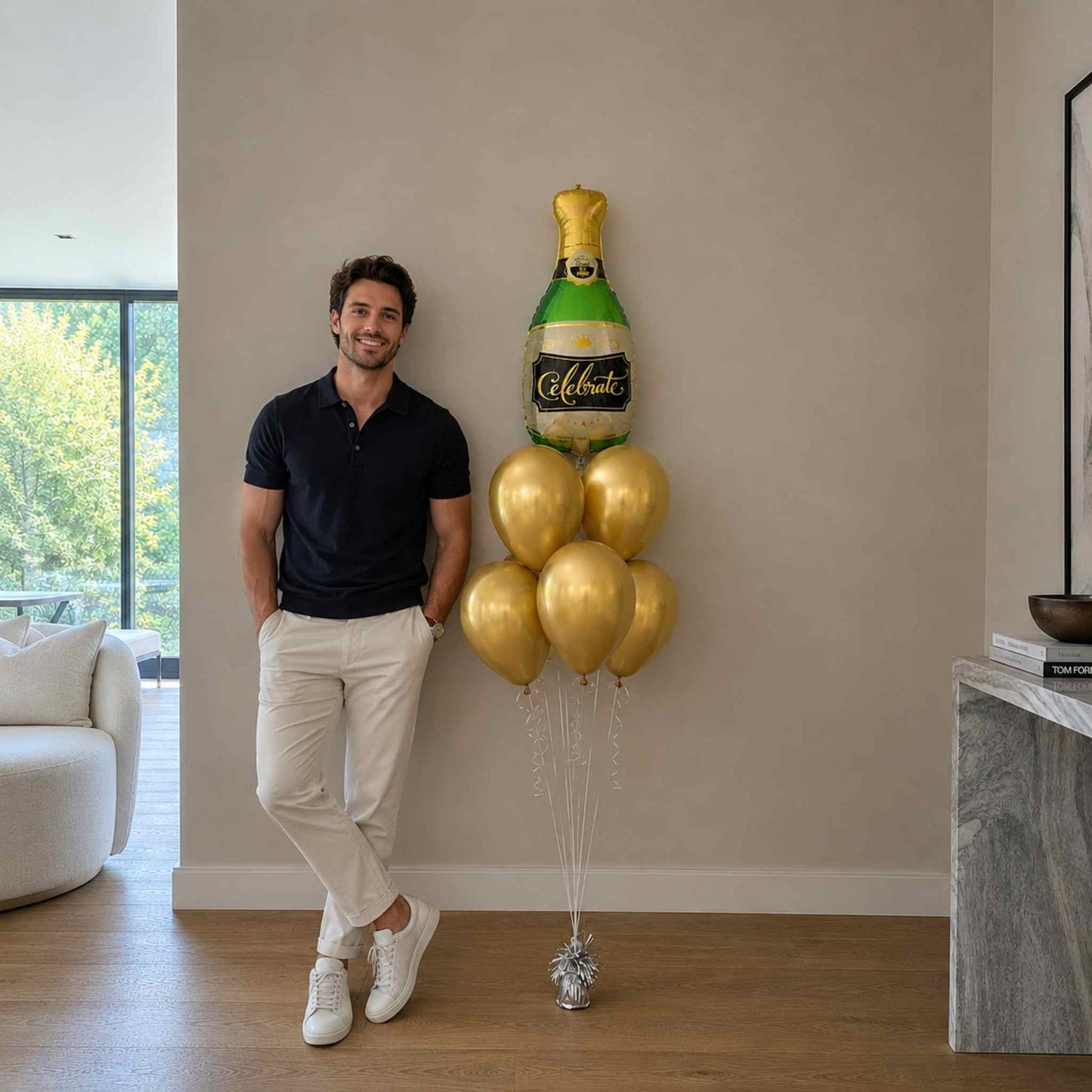 Man standing next to a large balloon shaped like a champagne bottle and bubbles in a living room.