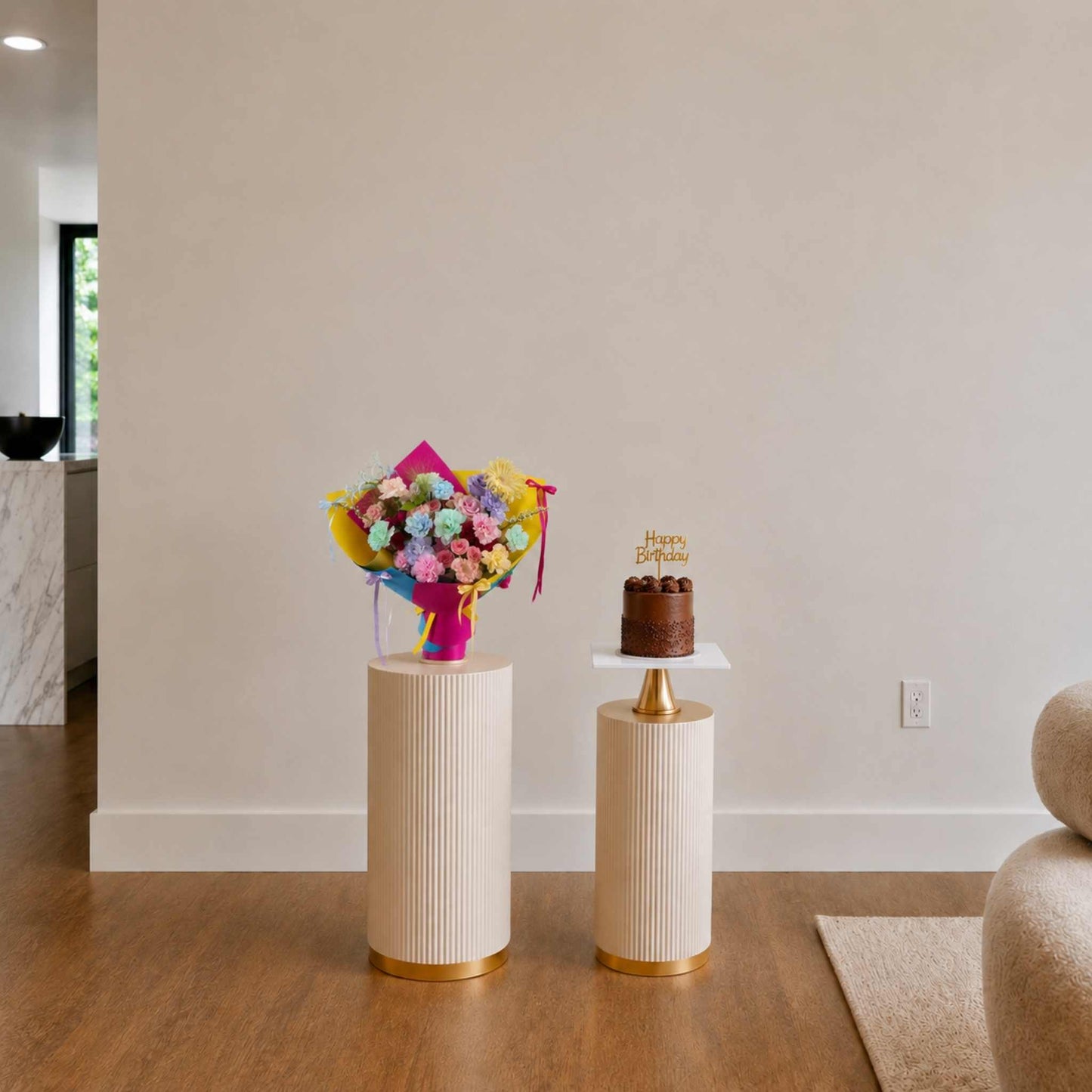 Bouquet of flowers and small chocolate cake on decorative stands against a plain wall.
