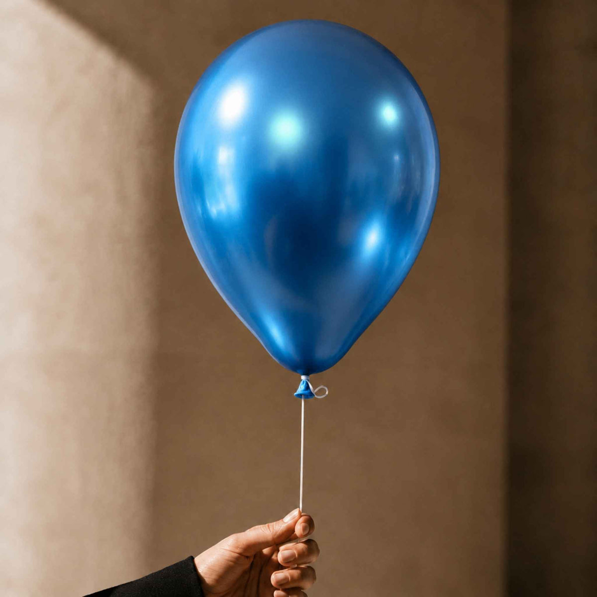 A shiny metallic blue balloon held by a hand against a warm beige wall background.