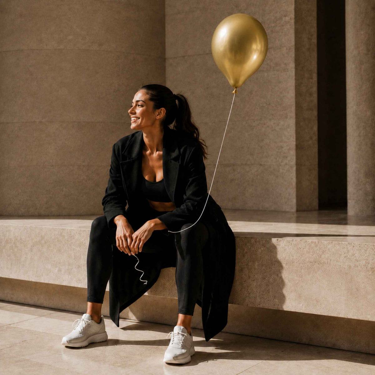 A woman sits on stone steps in a sunlit architectural setting, wearing a black blazer, black crop top, black leggings, and white sneakers, with a metallic gold balloon floating beside her.