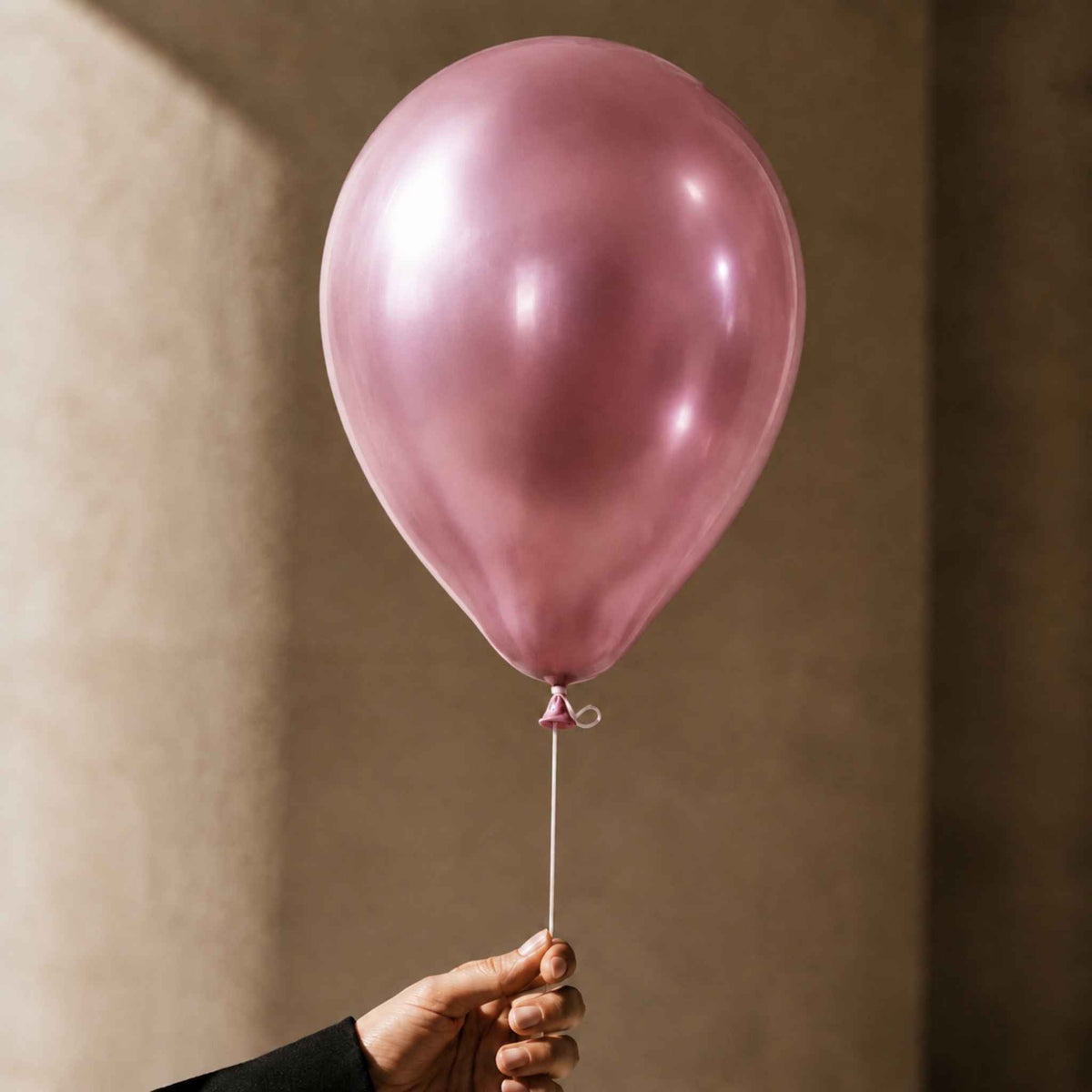 A single metallic dusty pink latex balloon held by a hand against a warm beige textured wall background.