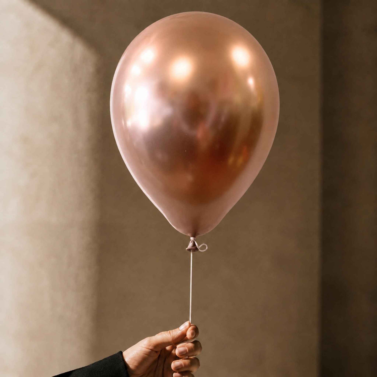 A metallic rose gold latex balloon held by a hand against a warm, textured beige background.