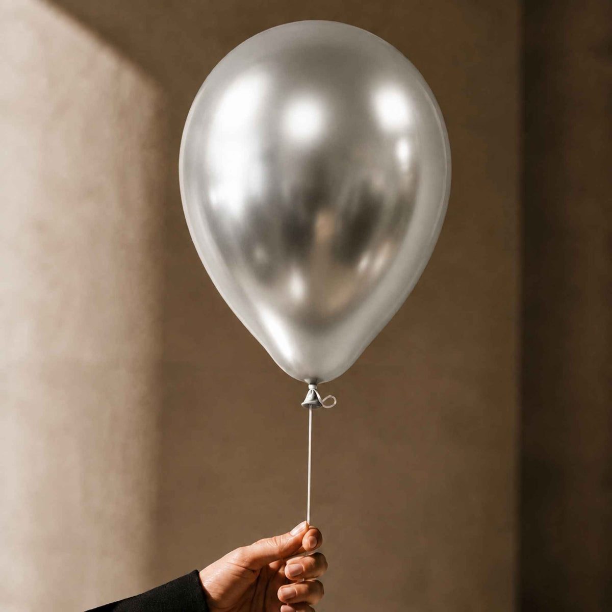 A metallic silver latex balloon held by a hand against a warm, textured beige wall.