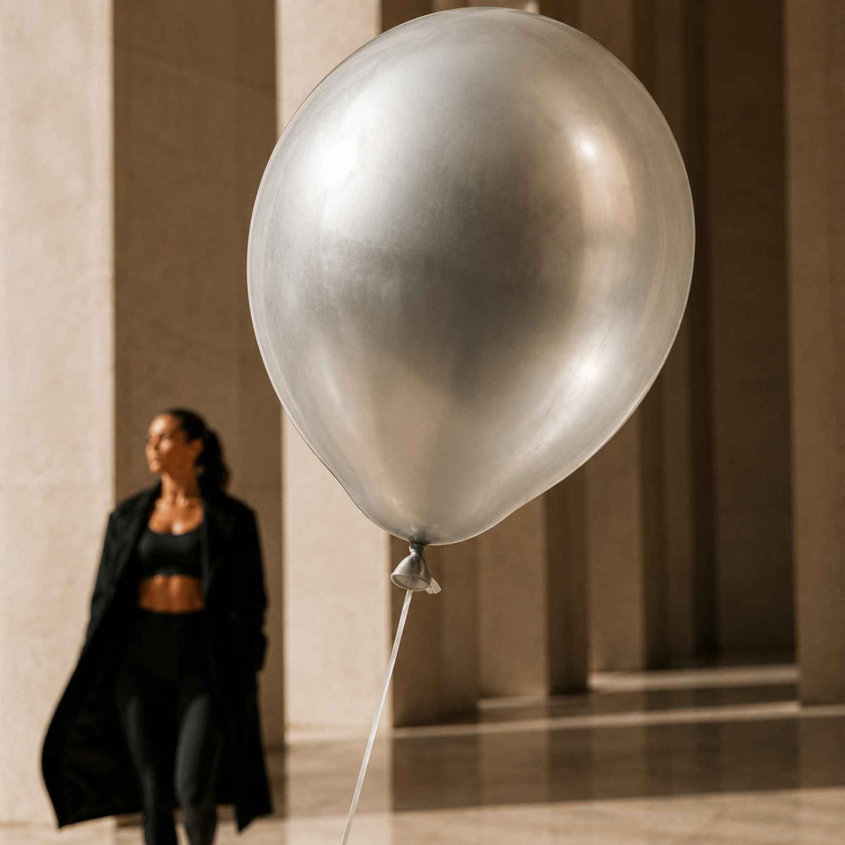A large metallic silver balloon dominates the foreground, with a blurred woman in a black outfit standing in the background near large stone columns.