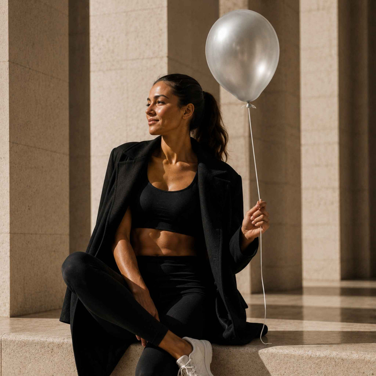 A woman in a black athletic set with an open black blazer sits on stone steps, holding a metallic silver balloon in a sunlit architectural setting.