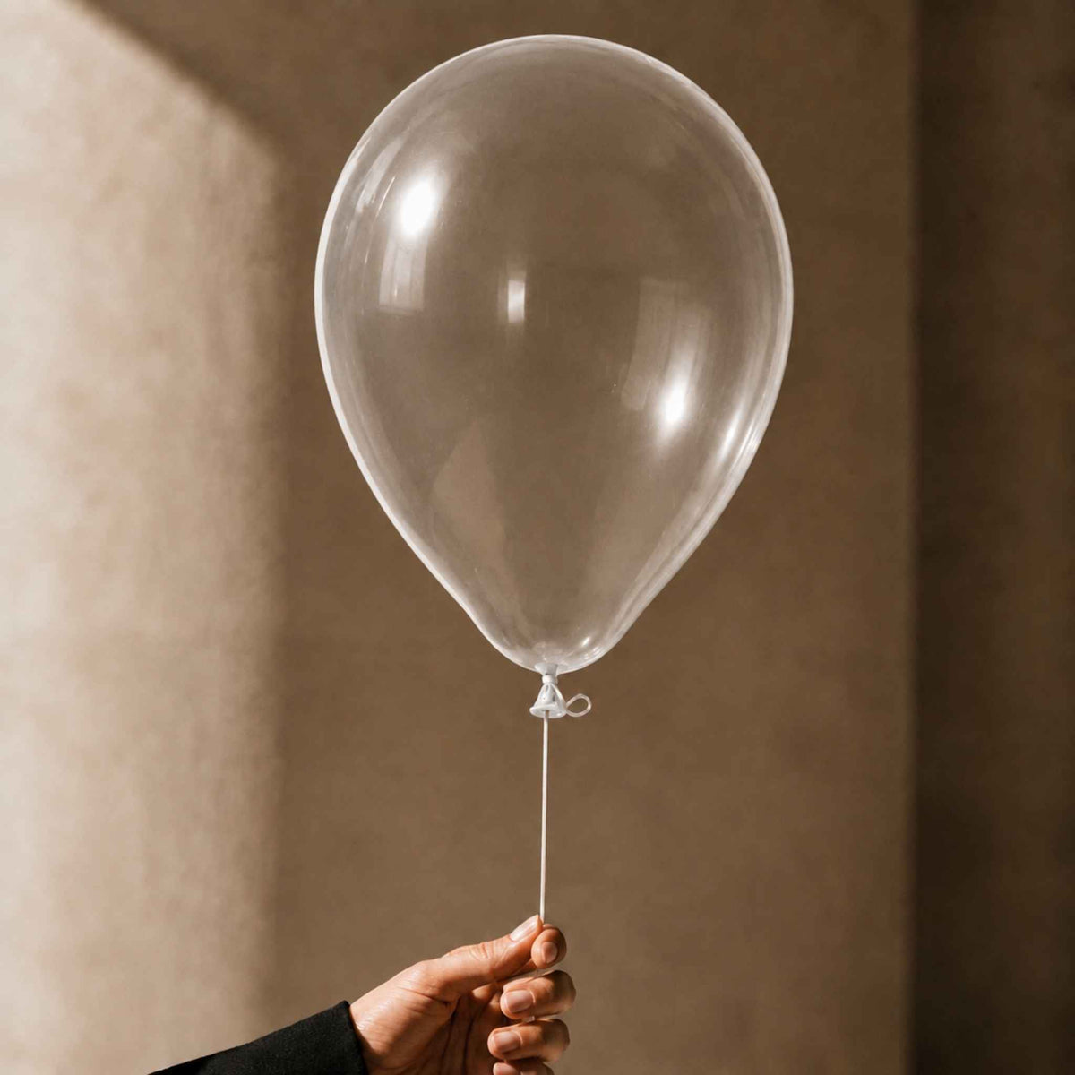 A hand holding a clear, round latex balloon with a white string against a warm, textured beige background.