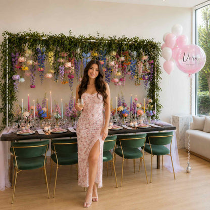 Woman standing in a room with a decorated table and floral arch, holding a glass.