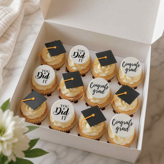 Box of cupcakes with graduation-themed decorations on a marble surface