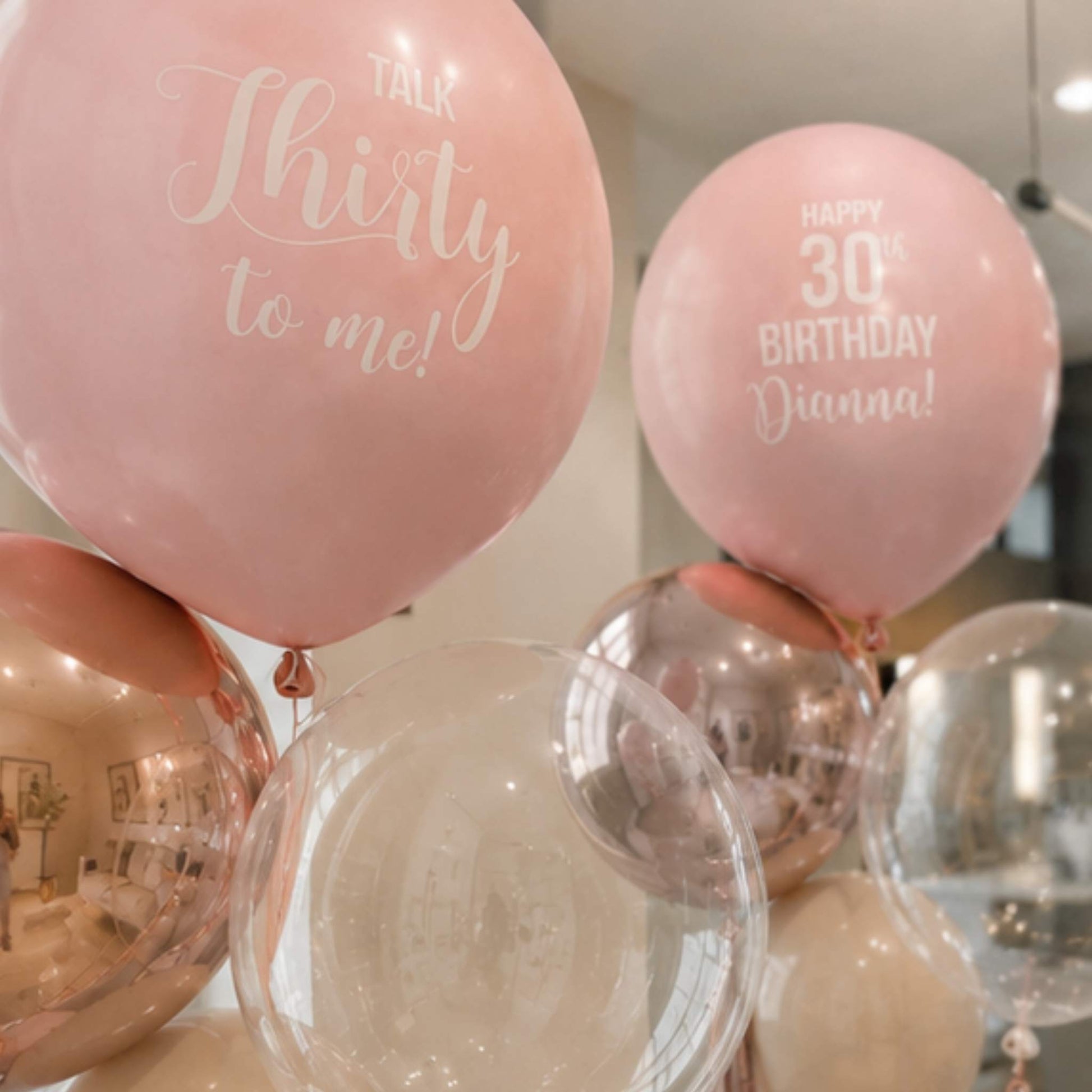 Pink latex balloons with white printed birthday text, including one that reads 'Talk Thirty to me!' and another that reads 'Happy 30th Birthday Dianna!', alongside clear and rose gold balloons in an indoor setting.