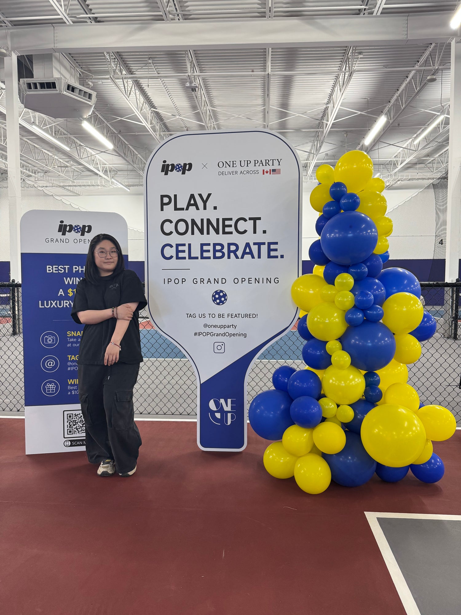 A woman stands indoors beside a large promotional sign that reads 'PLAY. CONNECT.