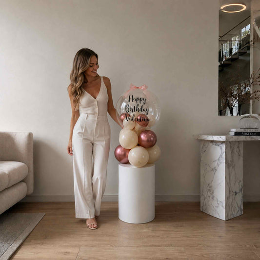 Woman in a white outfit standing next to a decorative balloon arrangement with 'Happy Birthday' text in a modern living room.