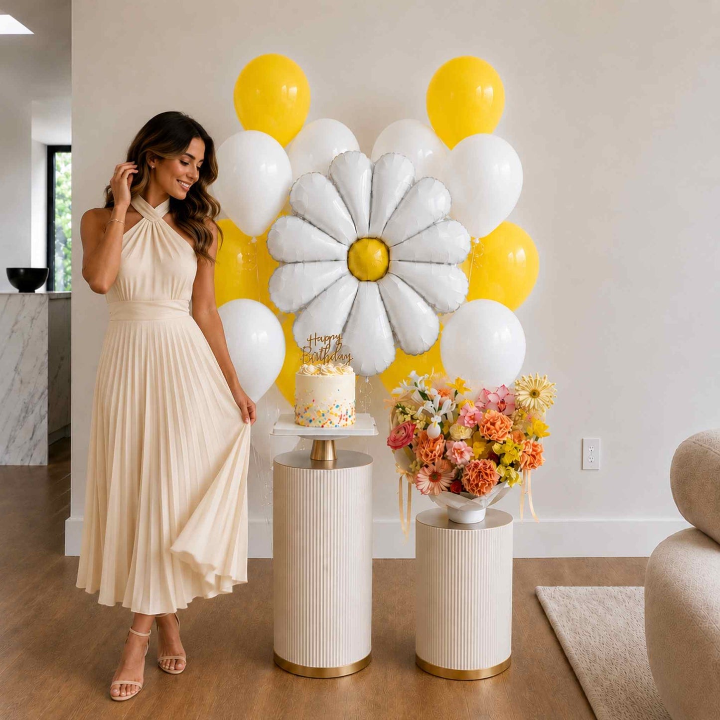 Woman in a beige dress standing next to a cake and floral arrangement with yellow, white, and gray balloons.