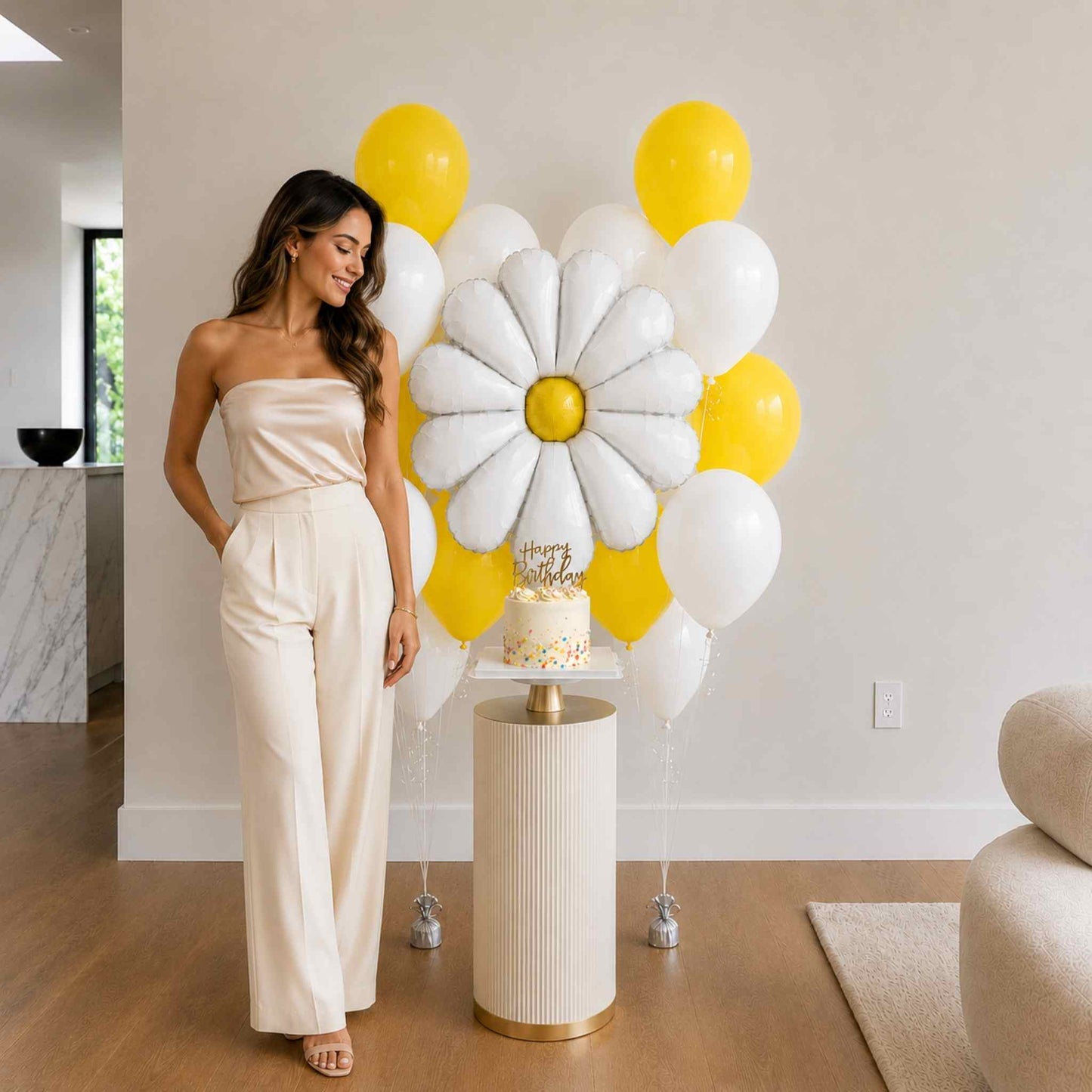 Woman in a beige outfit standing next to a floral balloon arrangement and cake in a room.