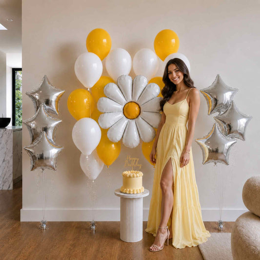 Woman in a yellow dress standing next to a cake and balloon arrangement in a room.