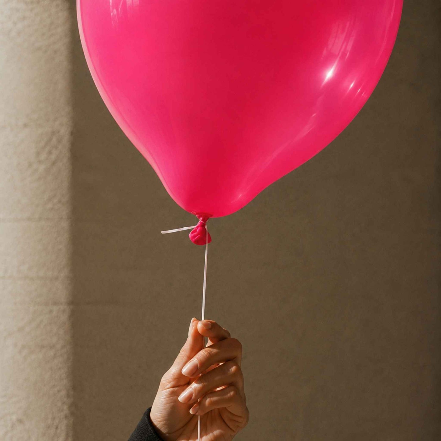 A bright pink balloon held by a hand against a neutral beige wall background.