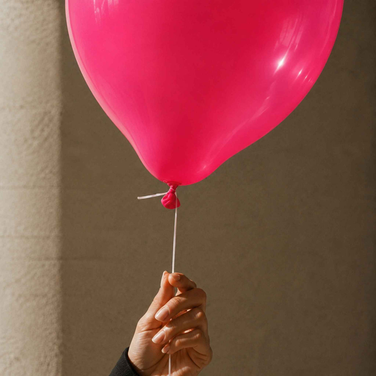 A bright pink balloon held by a hand against a neutral beige wall background.