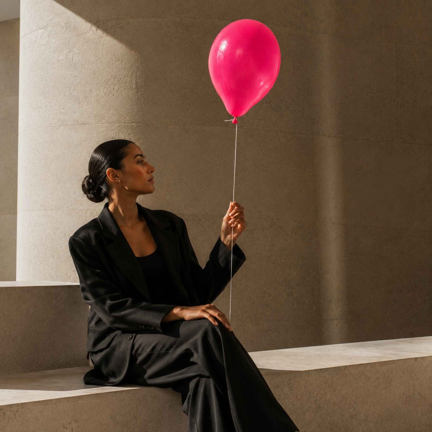 A woman in a black blazer and trousers sits on a concrete bench, looking up at a bright pink balloon she is holding by its string in a minimalist, sunlit architectural setting.