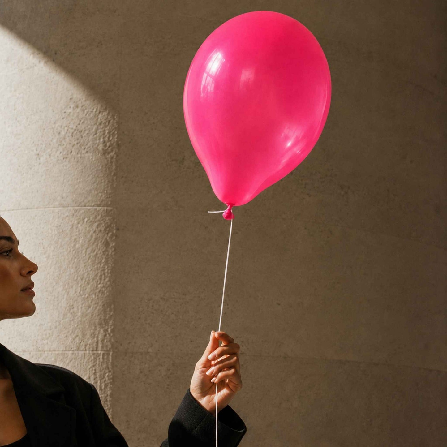 A person in a black blazer holds a bright pink balloon against a textured beige wall with dramatic side lighting.