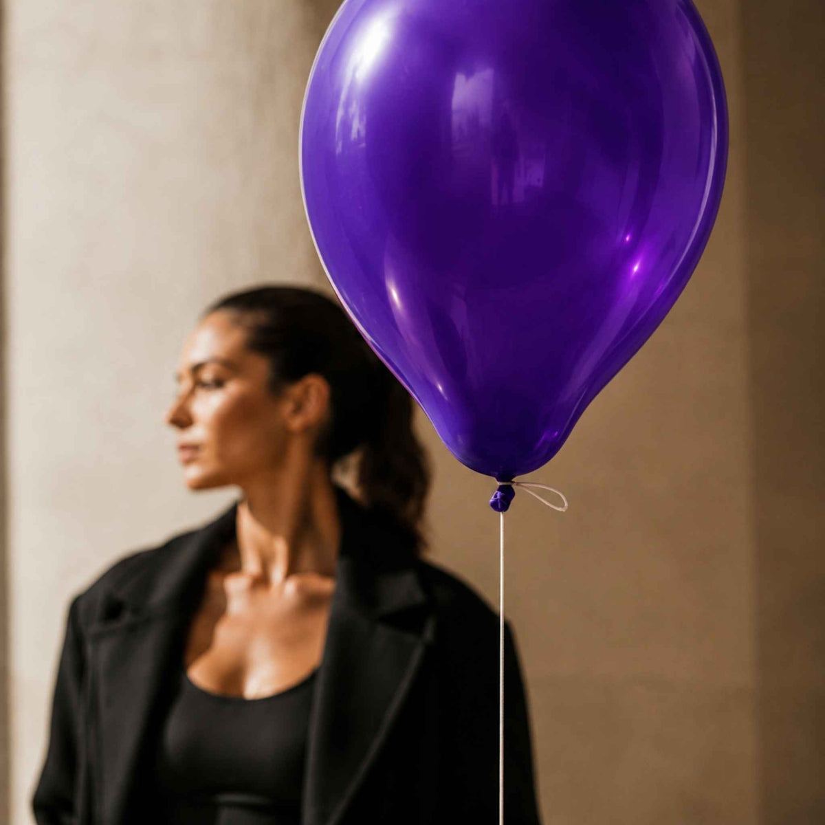 A large shiny purple balloon in the foreground with a blurred woman wearing a black blazer and black top in the background against a beige wall.