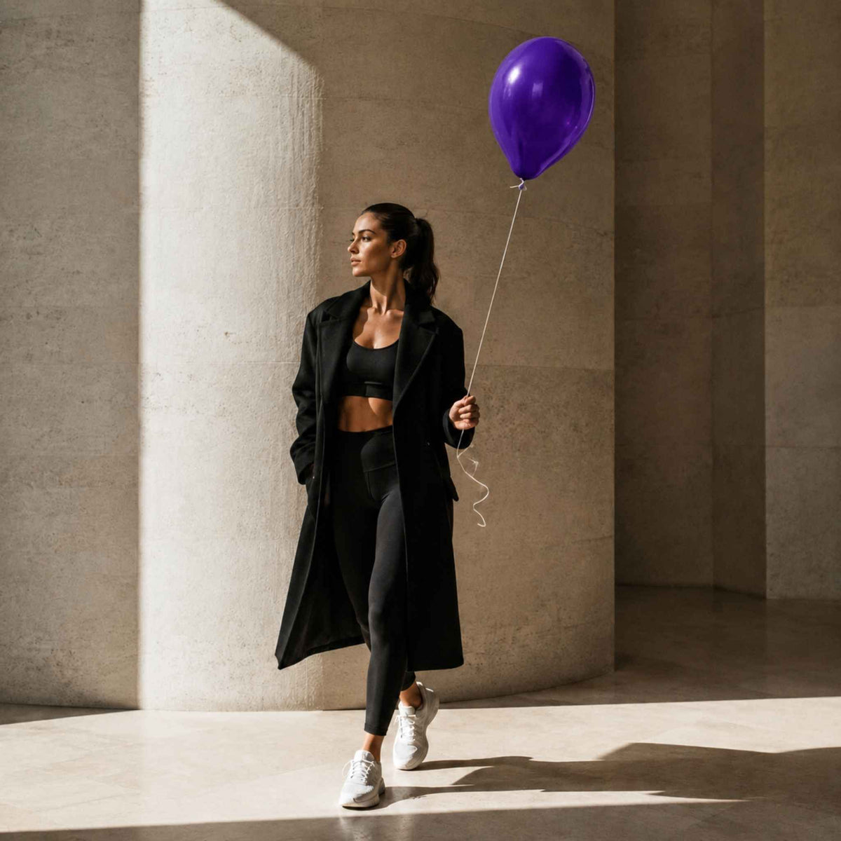 A woman in a black activewear set with a long black coat walks through a sunlit concrete corridor while holding a purple balloon.