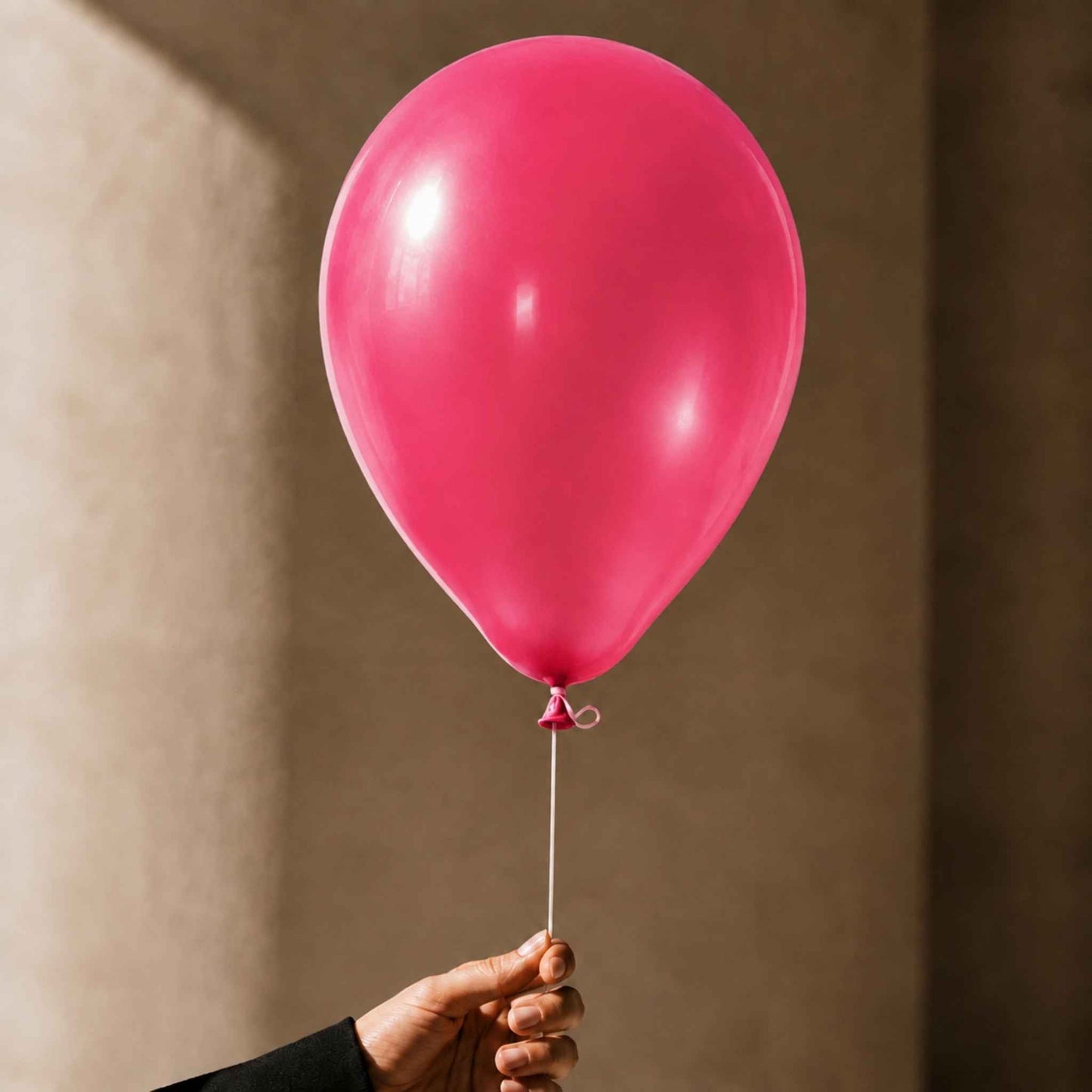 A single bright pink balloon held by a hand against a warm beige wall background.