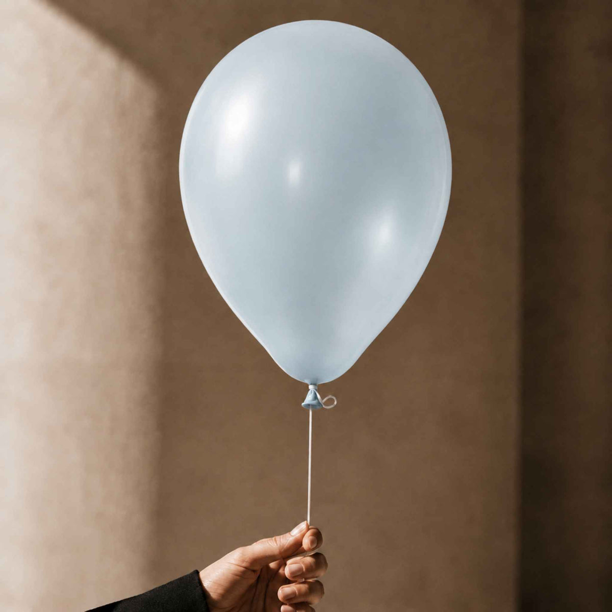A single pale blue latex balloon held by a hand against a warm beige wall with soft sunlight and shadows.