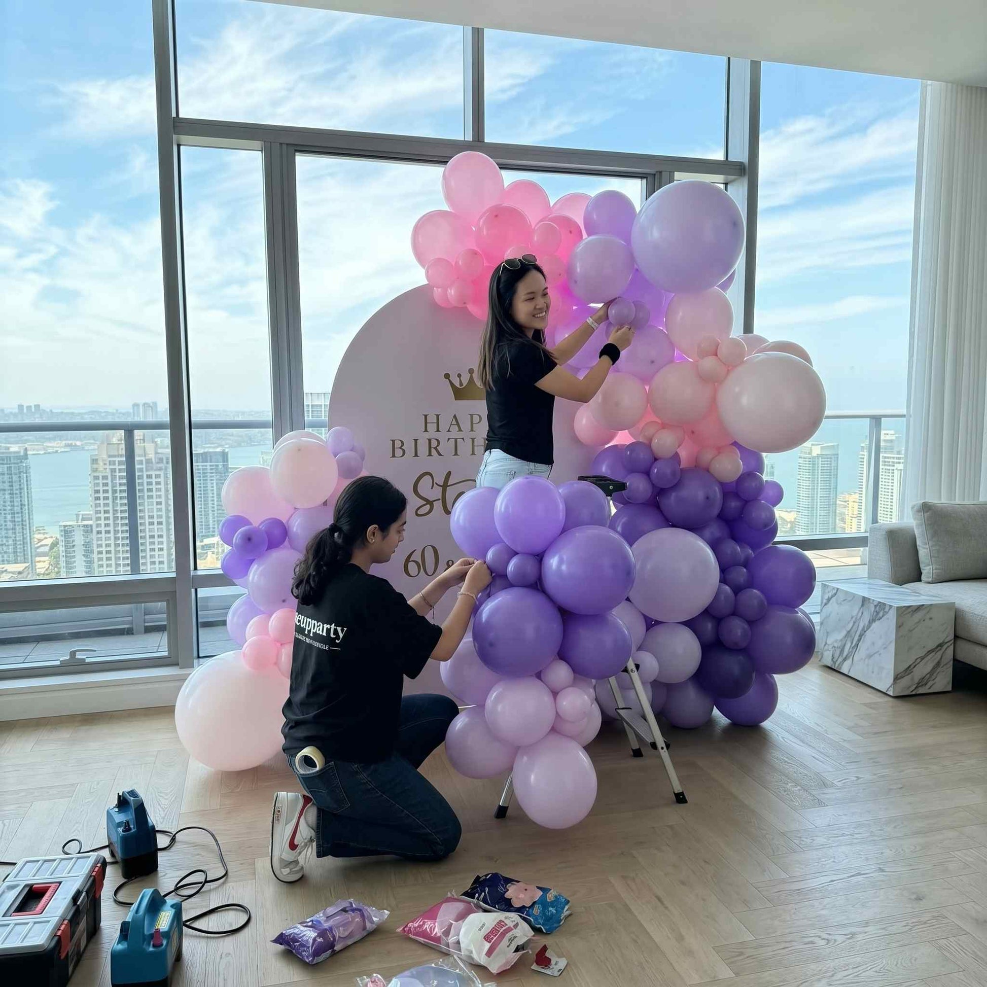 Balloon arch with 'Happy Birthday Stella 60 & Fabulous' sign in a room with large windows two people setting up