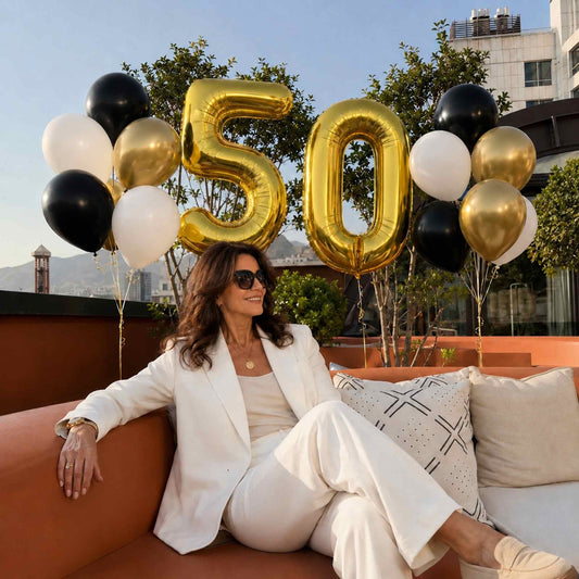 A woman in a white blazer and matching pants sits on an outdoor sofa with a large gold '50' balloon decoration and black, white, and gold balloons behind her.