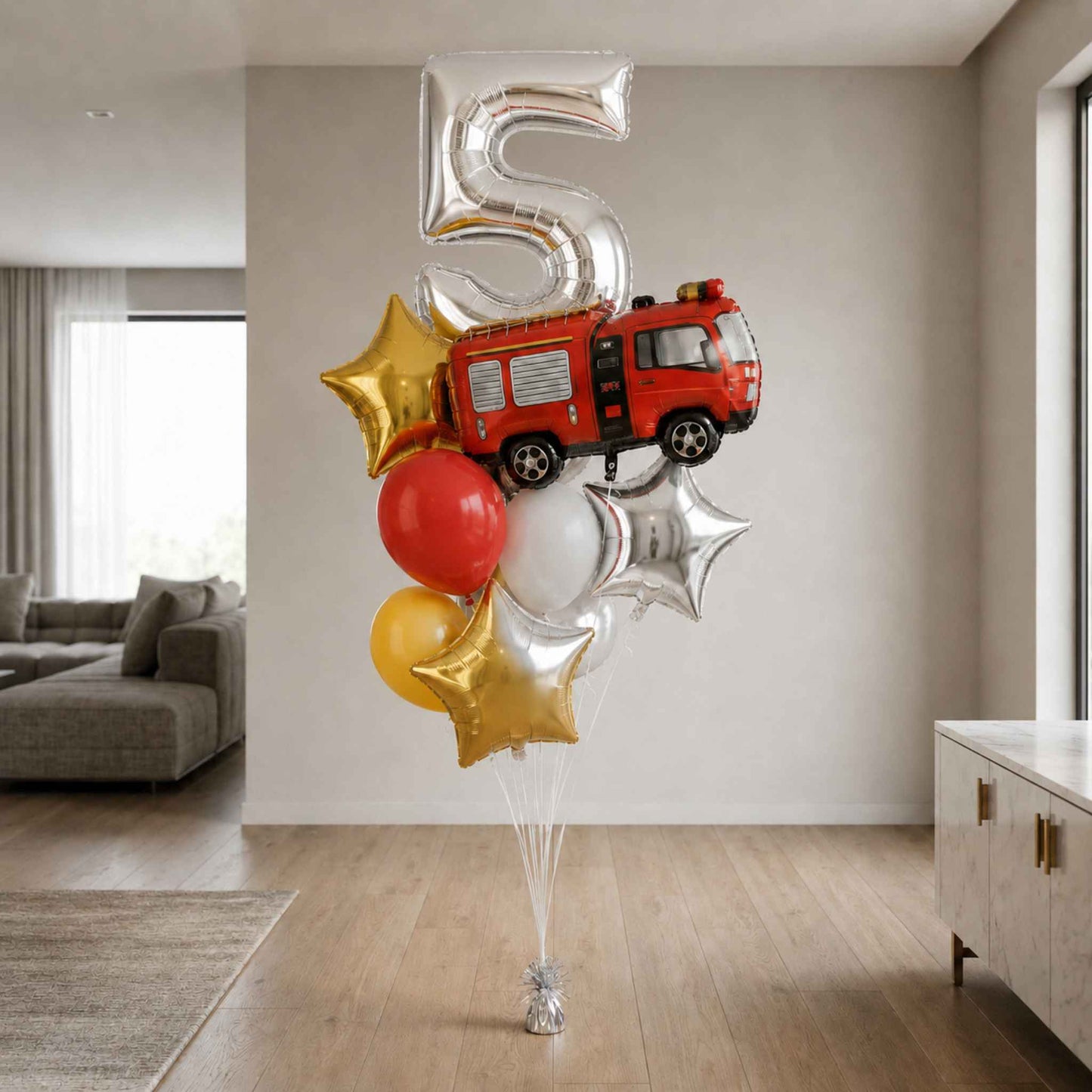 A balloon bouquet shaped like a number 5 birthday celebration, featuring a red fire truck, gold and silver star balloons, and a red and white balloon, standing on a floor in a modern living room.