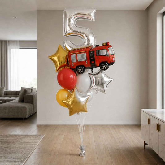 A balloon bouquet shaped like a number 5 birthday celebration, featuring a red fire truck, gold and silver star balloons, and a red and white balloon, standing on a floor in a modern living room.