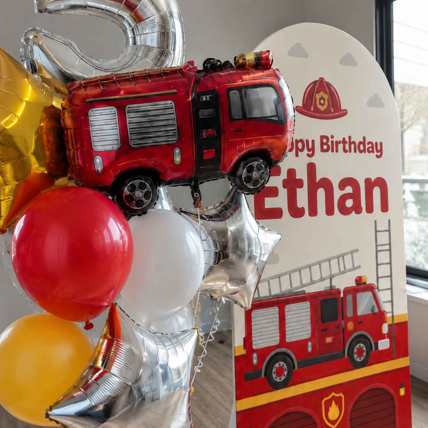 A personalized firefighter-themed birthday balloon bouquet with a red foil fire truck balloon, red, white, yellow, and silver balloons, and a matching custom 'Happy Birthday Ethan' sign featuring a fire truck illustration.