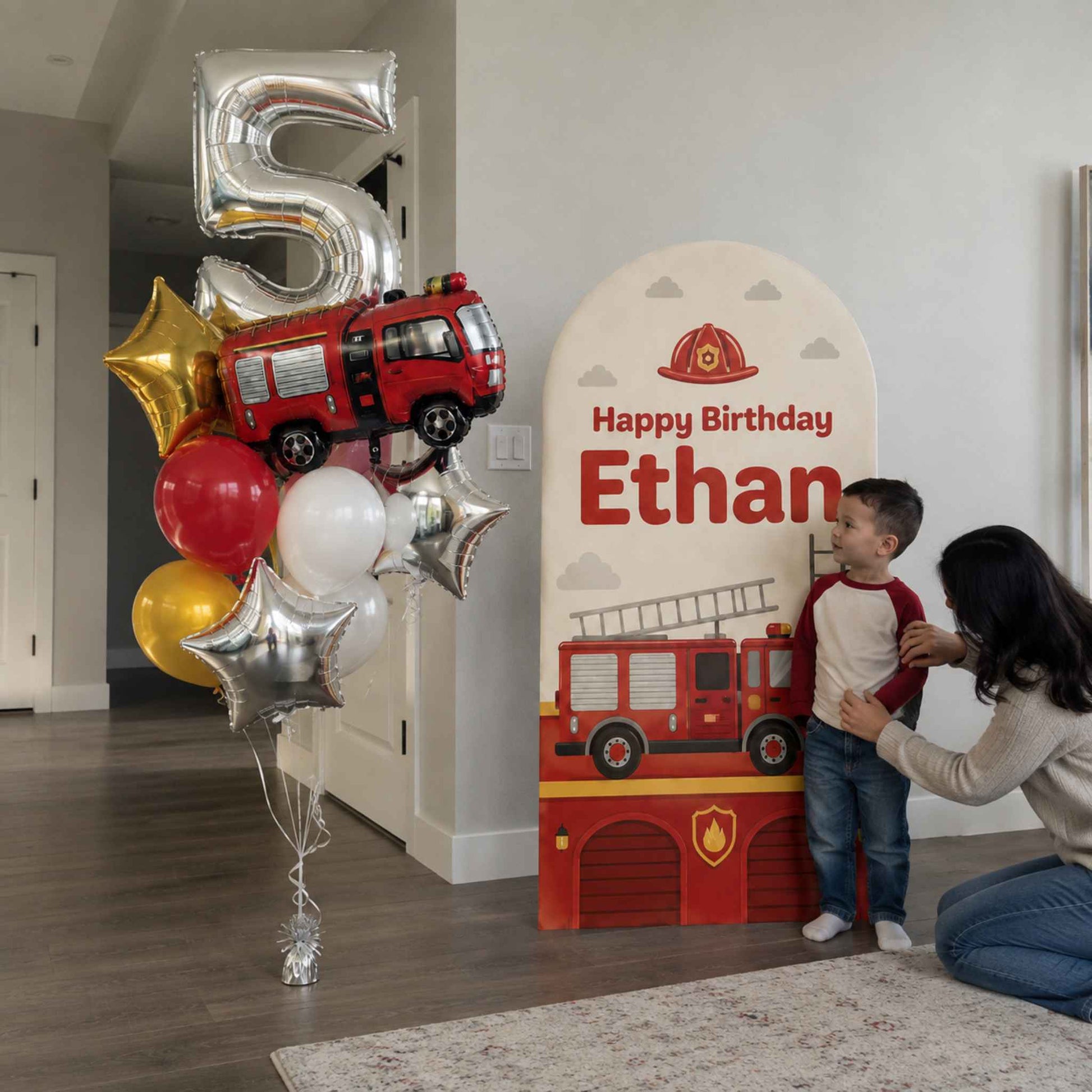 A child birthday party setup in a home interior, featuring a large metallic number 5 balloon cluster with a red fire truck, plus a custom fire station themed backdrop with the text 'Happy Birthday Ethan' and a woman adjusting a young boy standing in front of it.