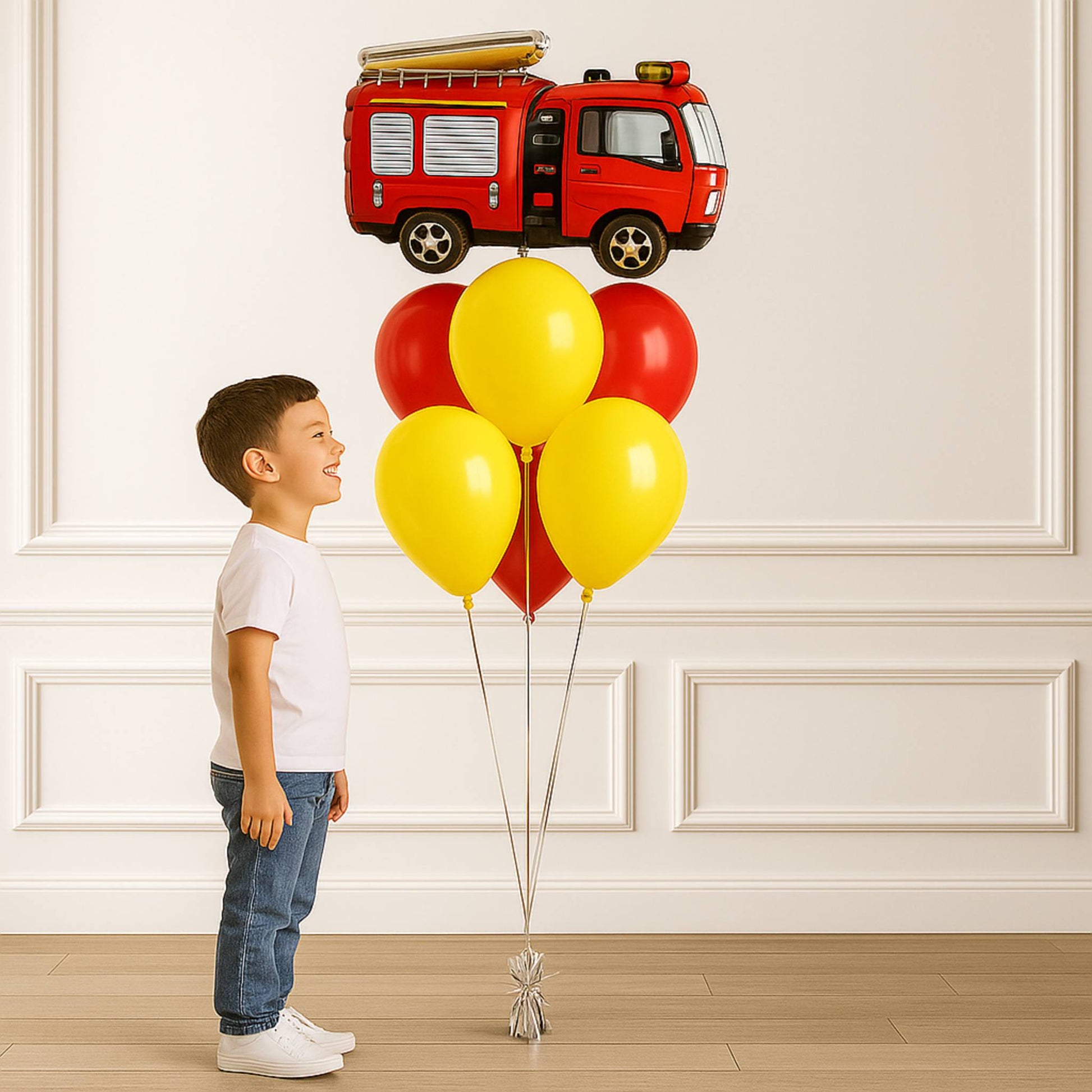 Child with red and yellow balloons shaped like a fire truck against a white paneled wall.