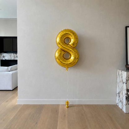Gold number 8 balloon on a plain wall in a room with a couch and TV.