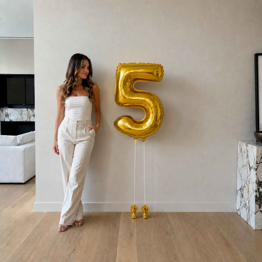 Woman in a white outfit standing next to a large gold number 5 balloon indoors.