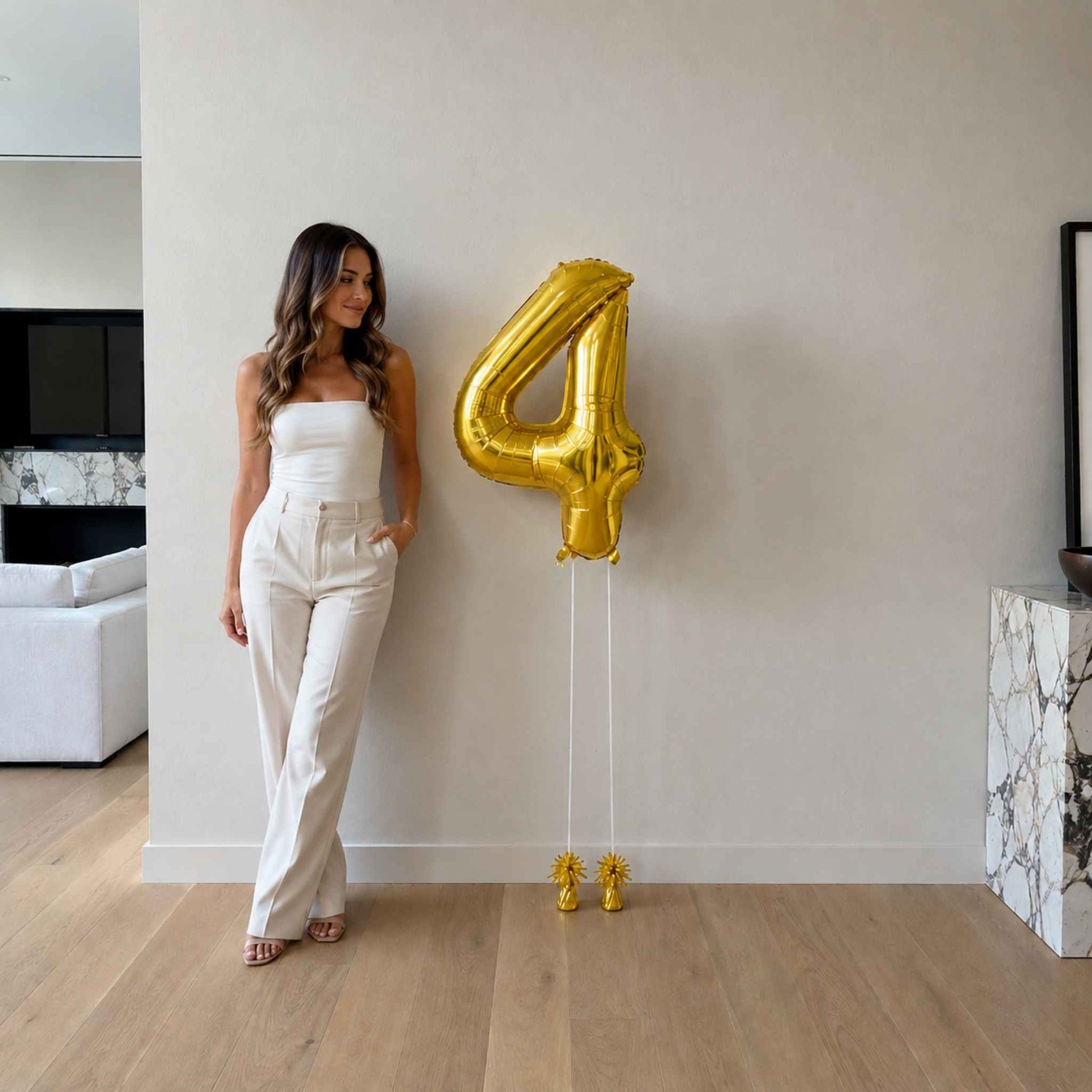 Woman in a white outfit standing next to a large gold number 4 balloon in a modern living room.
