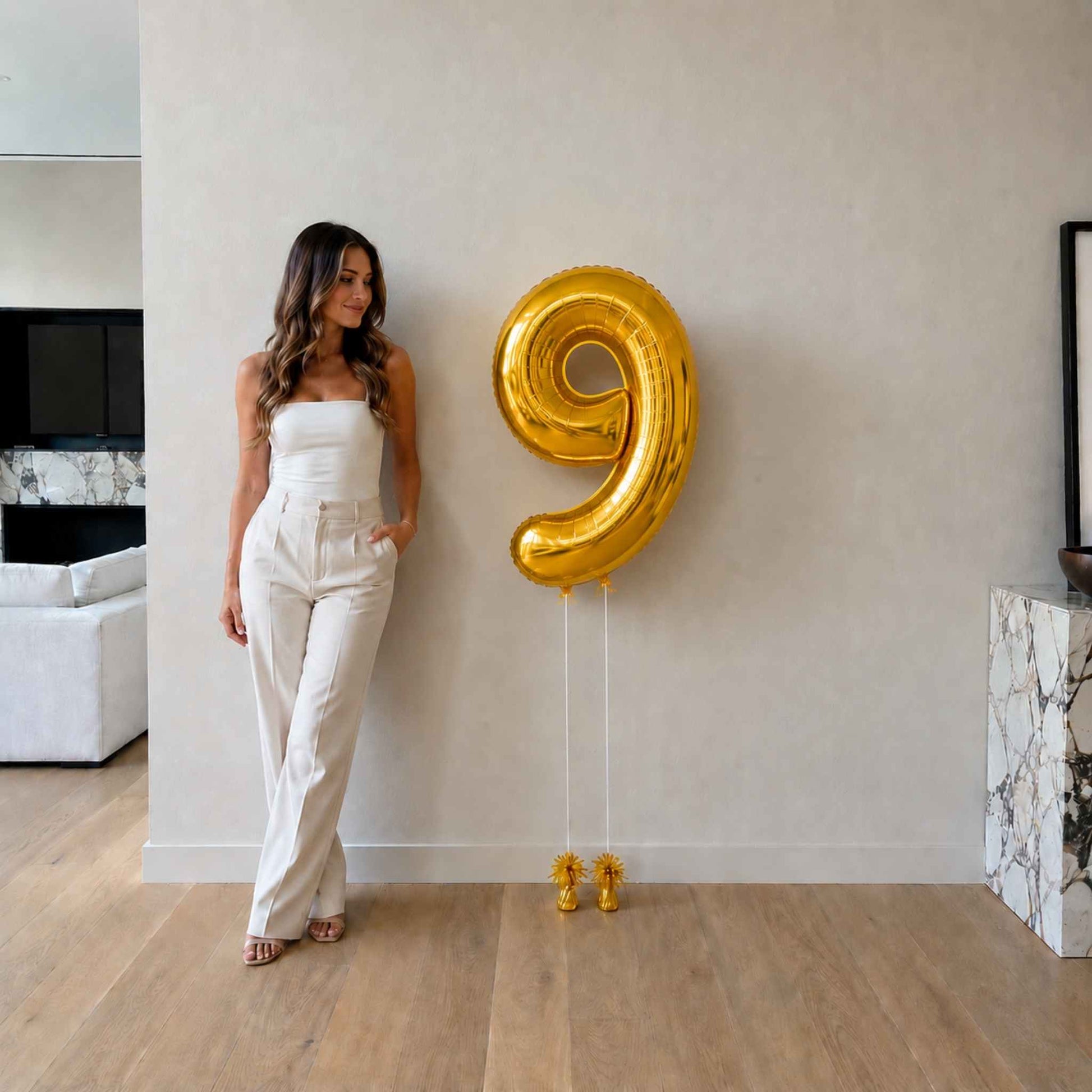 Woman standing next to a large gold number 9 balloon in a room with a white wall and wooden floor.