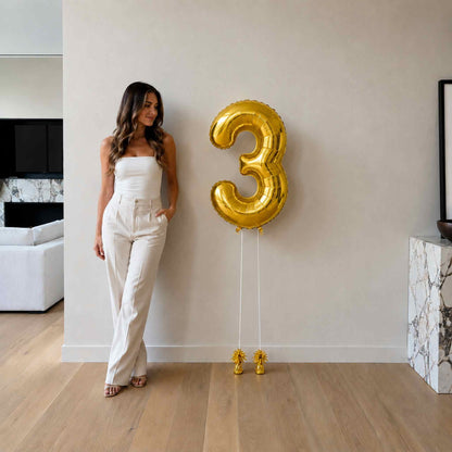Woman standing next to a large gold number three balloon in a modern living room.
