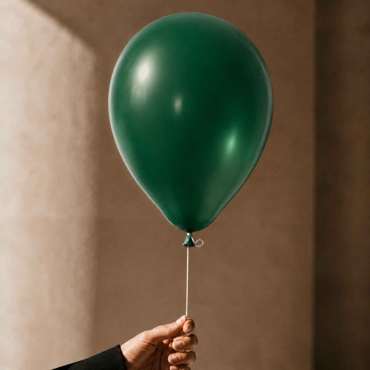 A single matte dark green balloon held by a hand against a warm beige textured wall.