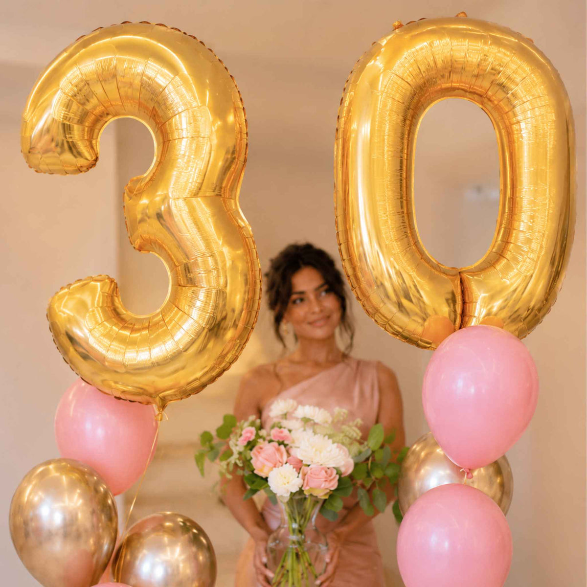 A woman holding a bouquet of pink and white flowers stands behind large metallic gold number 30 foil balloons, with smaller pink and gold balloons arranged around her in an indoor setting.