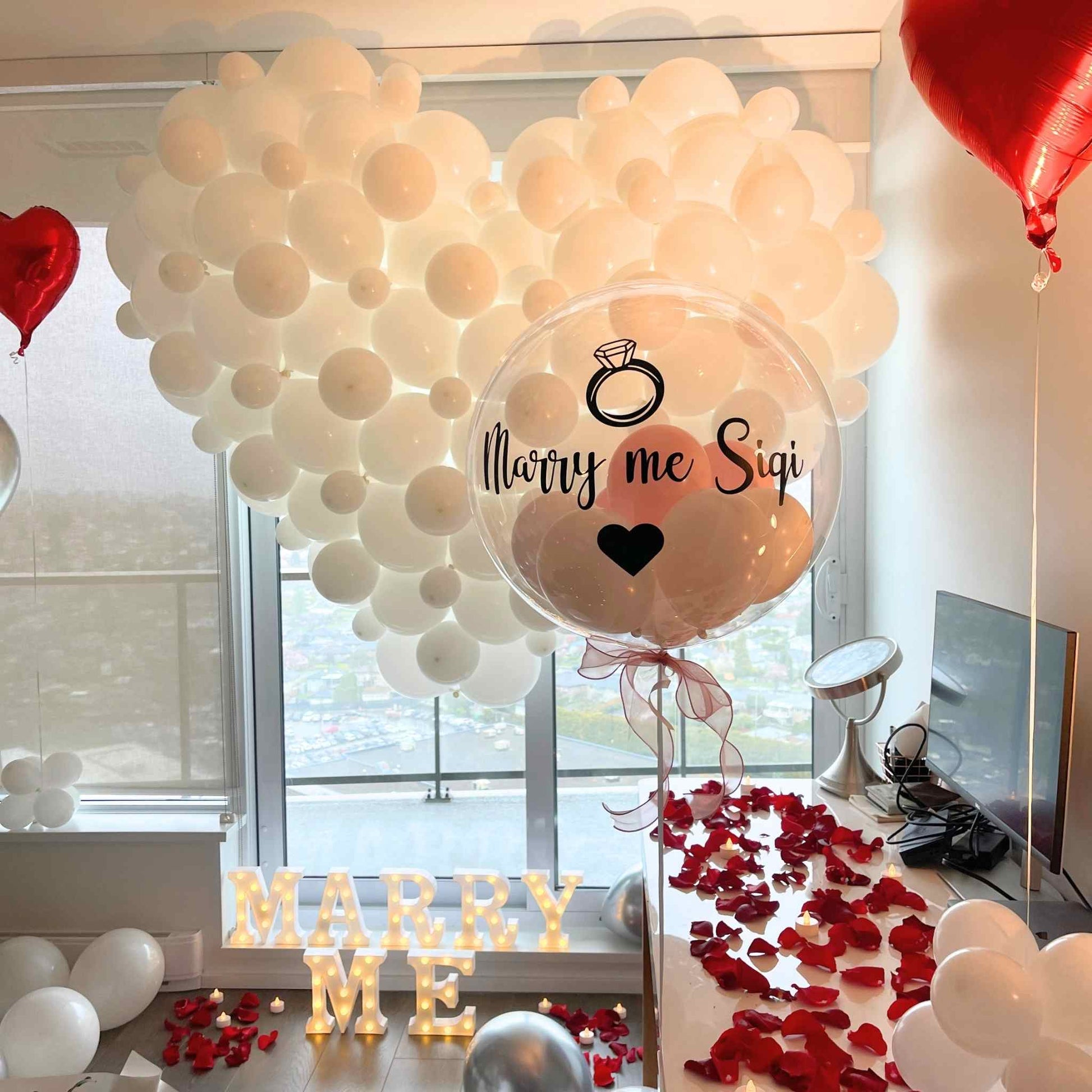 Heart-shaped balloon with 'Marry me Sigi' text in a room with balloons and rose petals.
