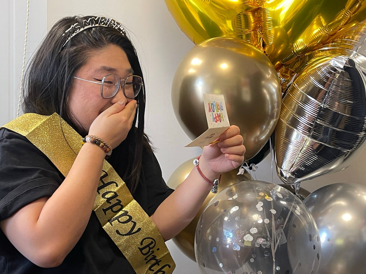 A person wearing a black t-shirt, a silver tiara, and a gold 'Happy Birthday' sash is blowing a kiss towards a small card held in their hand, with gold and silver balloons in the background.