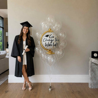 Graduate in cap and gown standing next to a large balloon with 'Go Change The World' text in a home setting.