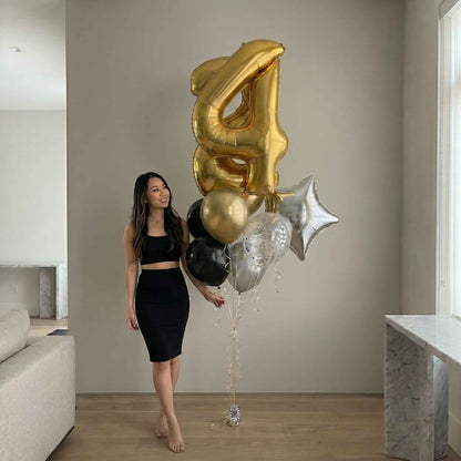 Woman holding a bouquet of gold, black, and silver balloons shaped like numbers in a modern living room.