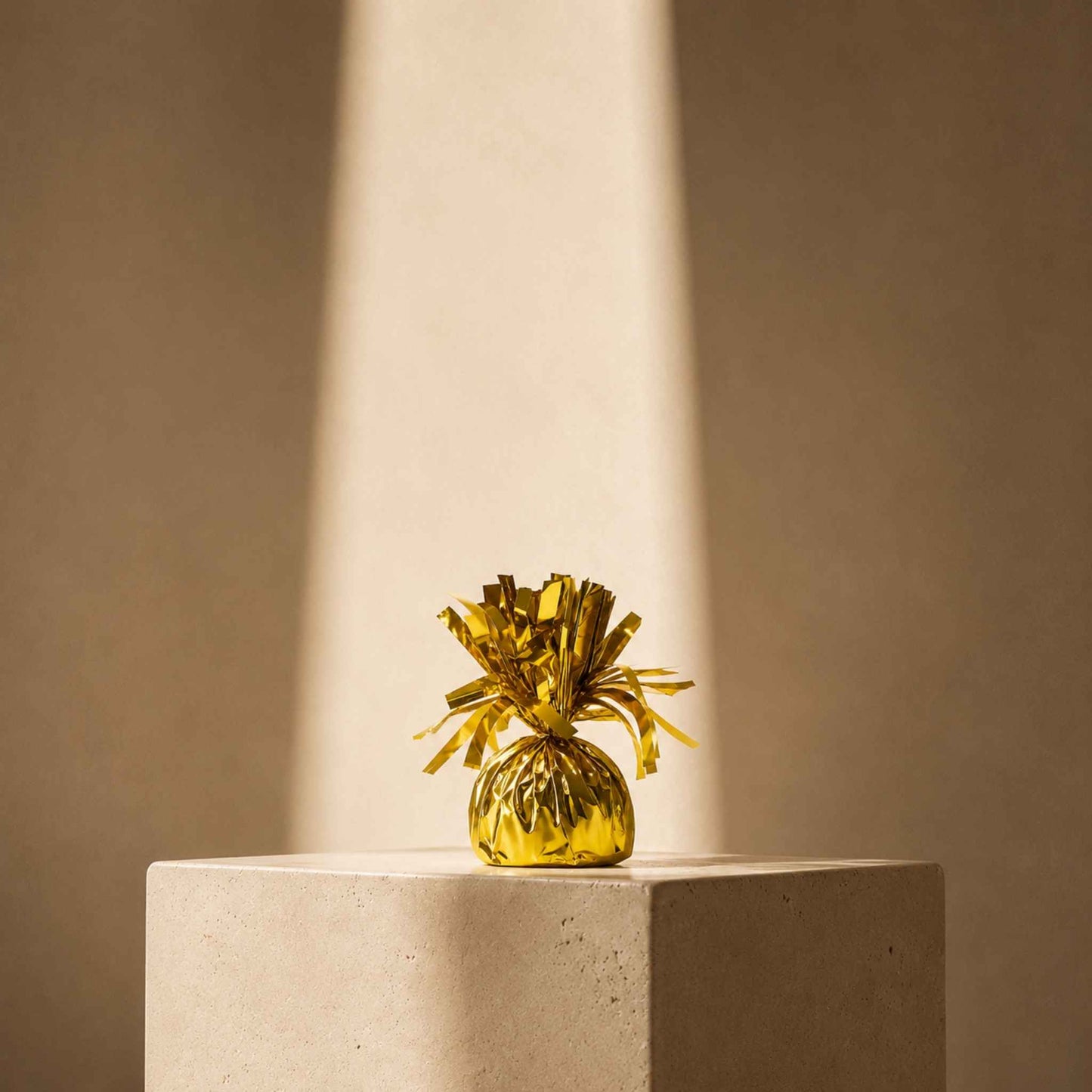 A small metallic gold tinsel pineapple decoration sits on a light beige stone pedestal, illuminated by a dramatic spotlight against a warm beige background.