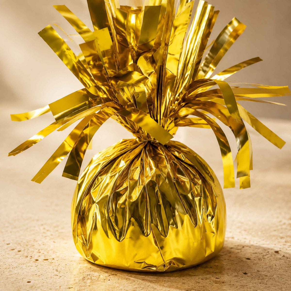 A shiny gold metallic foil-wrapped ornament or decorative bag with a tinsel-like fringe, photographed on a warm beige surface against a softly lit neutral background.
