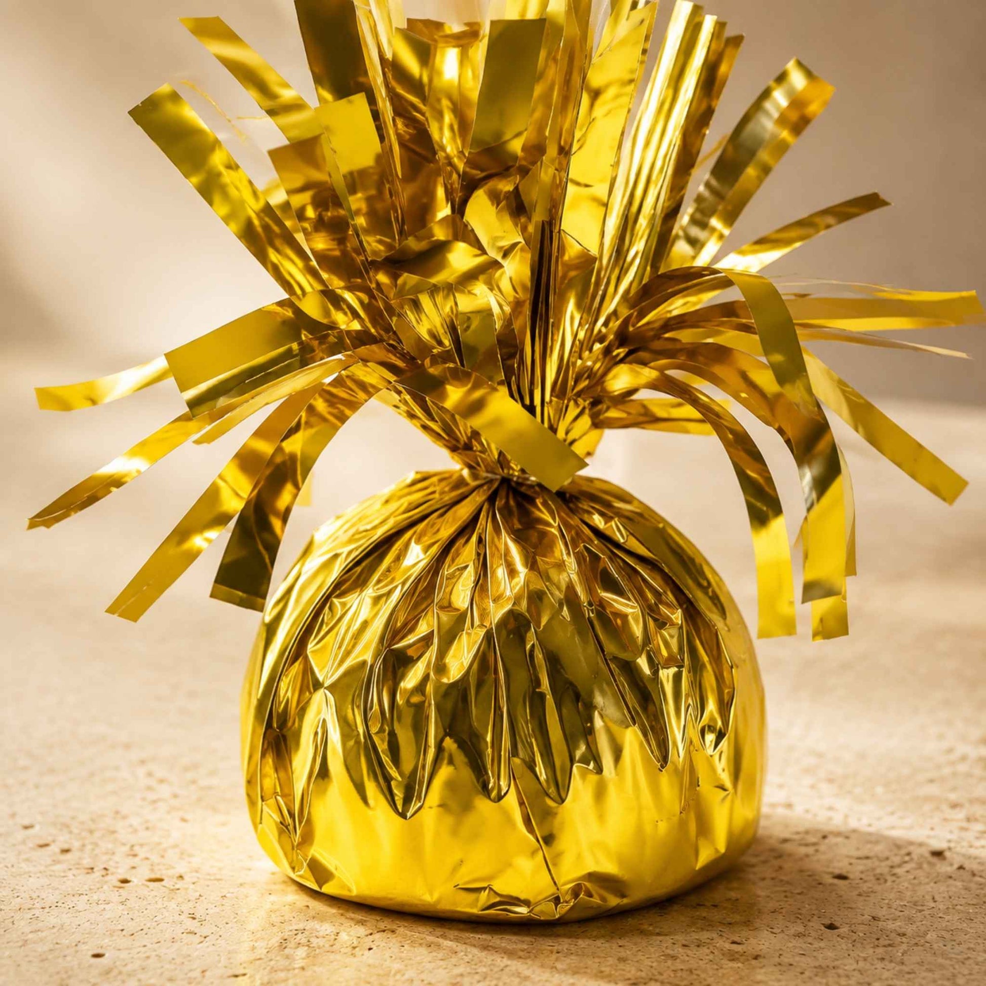 A shiny gold metallic foil-wrapped ornament or decorative bag with a tinsel-like fringe, photographed on a warm beige surface against a softly lit neutral background.